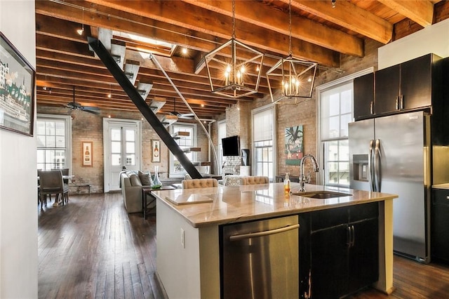 kitchen with brick wall, appliances with stainless steel finishes, pendant lighting, sink, and a kitchen island with sink