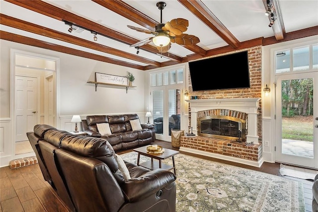 living room with track lighting, wood finished floors, ceiling fan, a brick fireplace, and beam ceiling