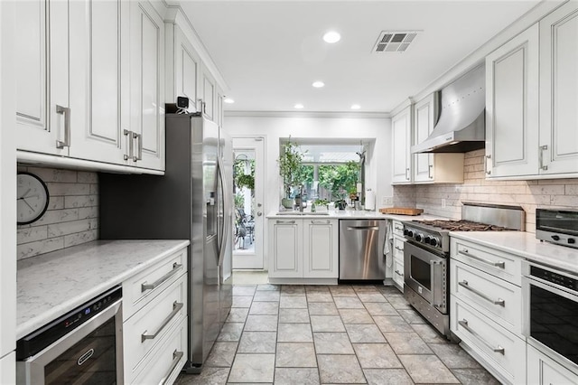 kitchen featuring stainless steel appliances, light stone countertops, white cabinets, beverage cooler, and wall chimney exhaust hood