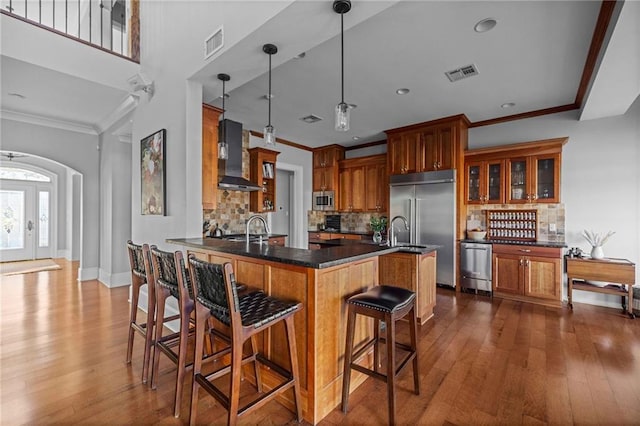 kitchen with a kitchen bar, hanging light fixtures, built in appliances, kitchen peninsula, and wall chimney exhaust hood