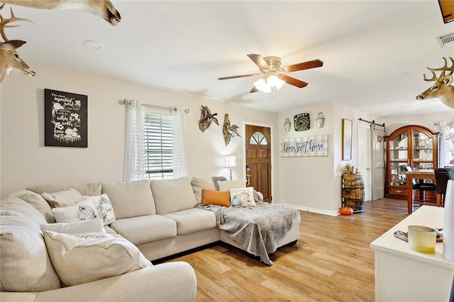 living room featuring ceiling fan, a barn door, visible vents, baseboards, and light wood finished floors