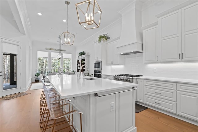 kitchen with sink, custom exhaust hood, white cabinetry, appliances with stainless steel finishes, and an island with sink