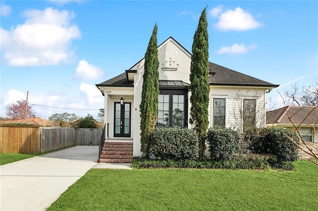 view of front facade featuring a front lawn, fence, roof with shingles, french doors, and driveway