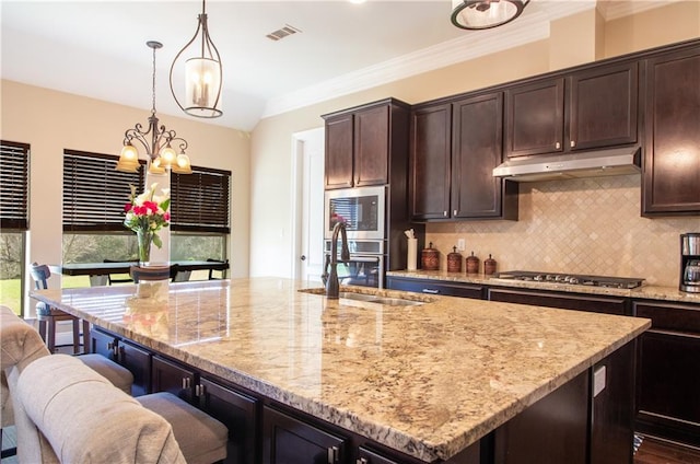 kitchen featuring a center island with sink, built in microwave, stainless steel gas stovetop, dark brown cabinetry, and decorative light fixtures