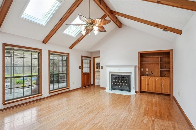 unfurnished living room with a skylight, light wood finished floors, a fireplace with flush hearth, ceiling fan, and beamed ceiling