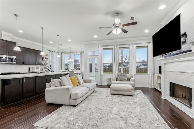 living area featuring baseboards, visible vents, dark wood finished floors, crown molding, and a stone fireplace