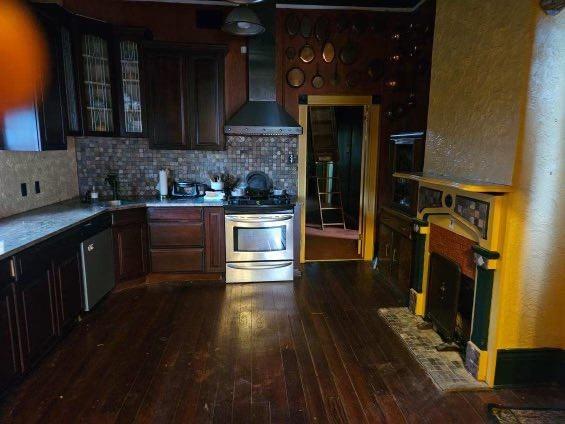 kitchen with stainless steel stove, decorative backsplash, dark wood-type flooring, dark brown cabinetry, and wall chimney exhaust hood