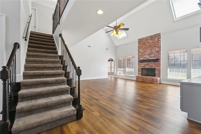 unfurnished living room with a skylight, a ceiling fan, dark wood-type flooring, a brick fireplace, and high vaulted ceiling