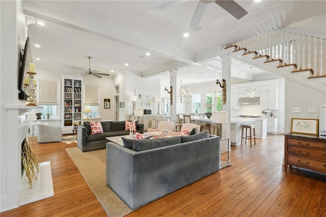 living area featuring a fireplace, beamed ceiling, light wood-style flooring, and ceiling fan with notable chandelier