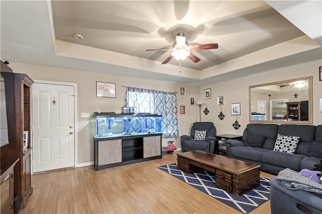 living area featuring ceiling fan, light wood-type flooring, a raised ceiling, and baseboards