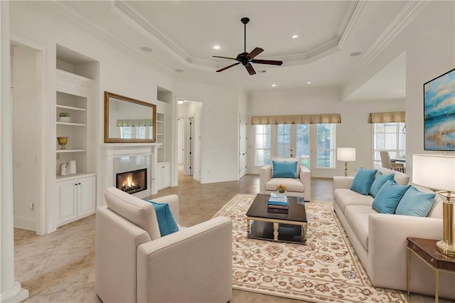 living room with a raised ceiling, a lit fireplace, crown molding, built in shelves, and recessed lighting