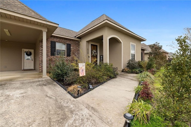 doorway to property featuring brick siding, roof with shingles, and stucco siding