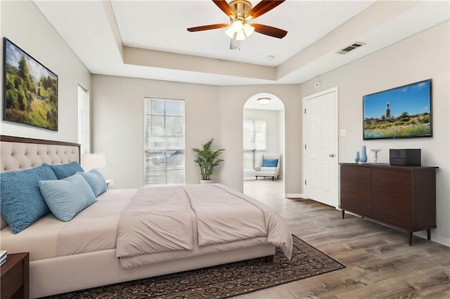 bedroom featuring arched walkways, visible vents, a raised ceiling, ceiling fan, and wood finished floors