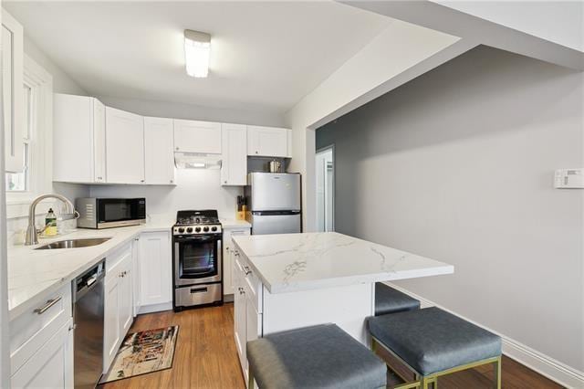 kitchen with a kitchen island, appliances with stainless steel finishes, light stone counters, under cabinet range hood, and a sink