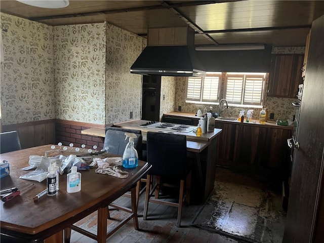 kitchen featuring dark brown cabinetry, wallpapered walls, light countertops, wall chimney range hood, and a sink