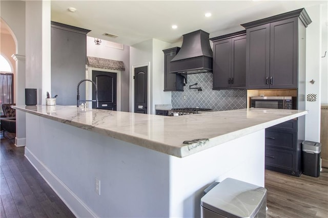 kitchen with arched walkways, decorative backsplash, light stone counters, dark wood-type flooring, and custom exhaust hood