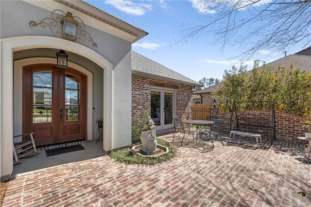 doorway to property with french doors, roof with shingles, brick siding, stucco siding, and fence