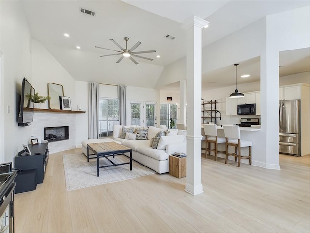 living area with ceiling fan, visible vents, light wood finished floors, a glass covered fireplace, and decorative columns