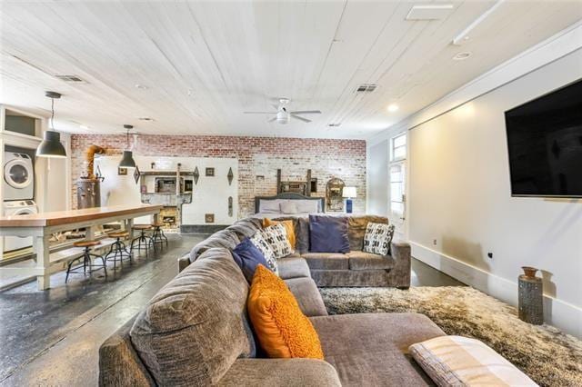 living room featuring finished concrete flooring, baseboards, stacked washer / dryer, brick wall, and ceiling fan