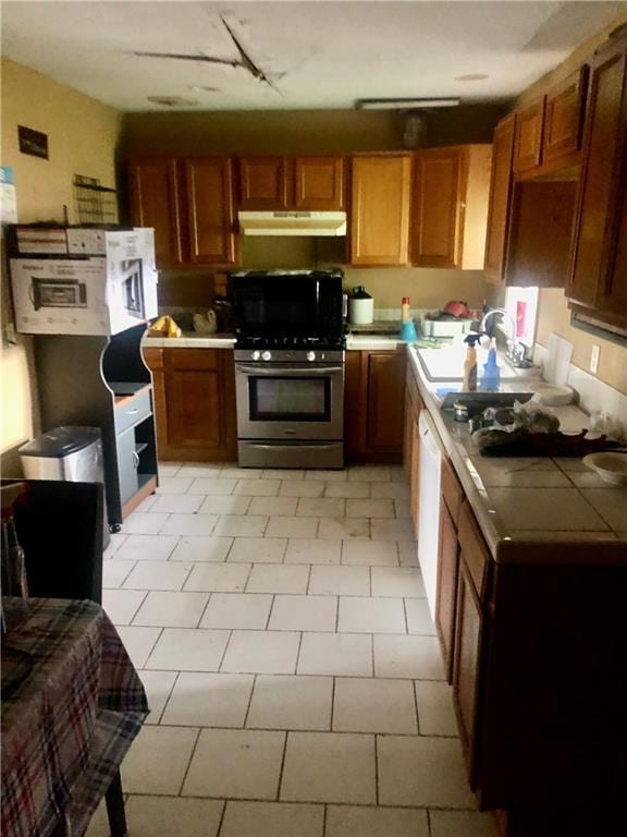 kitchen with brown cabinetry, tile countertops, under cabinet range hood, a sink, and gas stove