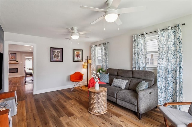 living area featuring a ceiling fan, a glass covered fireplace, baseboards, and wood finished floors