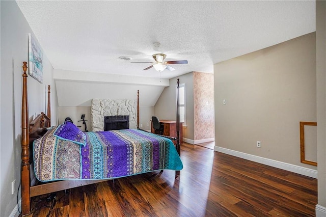 bedroom with vaulted ceiling, a stone fireplace, a textured ceiling, wood finished floors, and baseboards