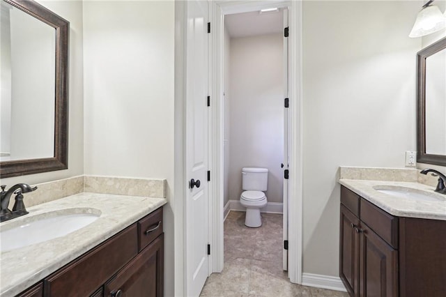 full bath featuring baseboards, two vanities, a sink, and toilet