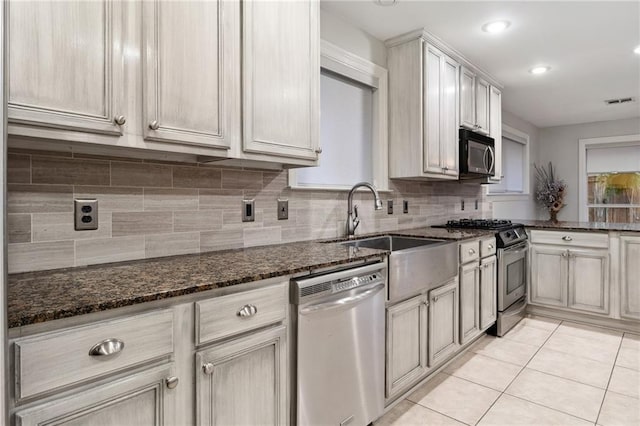 kitchen with light tile patterned flooring, stainless steel appliances, a sink, backsplash, and dark stone counters