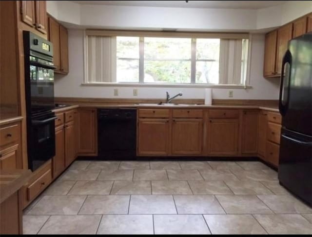 kitchen with brown cabinetry, light tile patterned floors, black appliances, and a sink