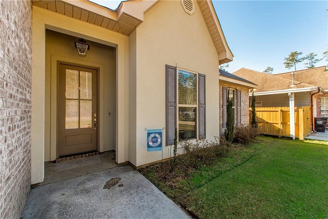 view of exterior entry featuring brick siding, a yard, fence, and stucco siding