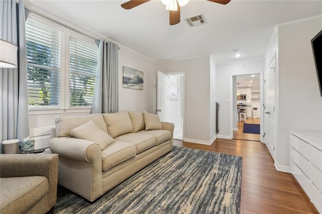 living room with dark wood-style flooring, a ceiling fan, visible vents, baseboards, and ornamental molding