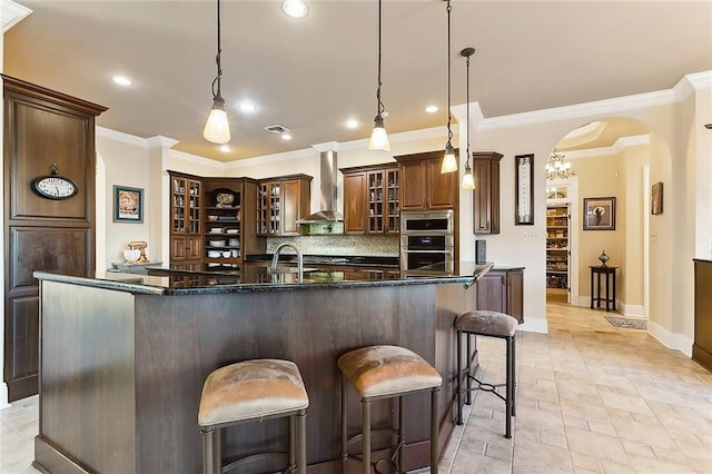 kitchen with arched walkways, visible vents, wall chimney range hood, stainless steel microwave, and a kitchen bar