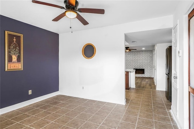 empty room with baseboards, brick wall, ceiling fan, tile patterned floors, and a brick fireplace