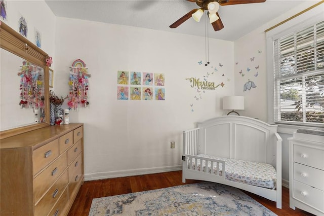 bedroom with a crib, a ceiling fan, baseboards, and dark wood-type flooring