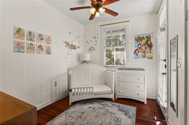 bedroom featuring a crib, ceiling fan, baseboards, and dark wood-style flooring