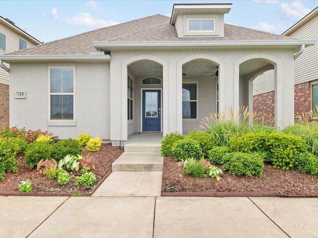 entrance to property with a shingled roof, a porch, a ceiling fan, and stucco siding