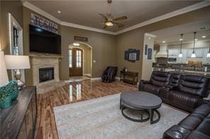 living room with arched walkways, a fireplace, ornamental molding, a ceiling fan, and wood finished floors