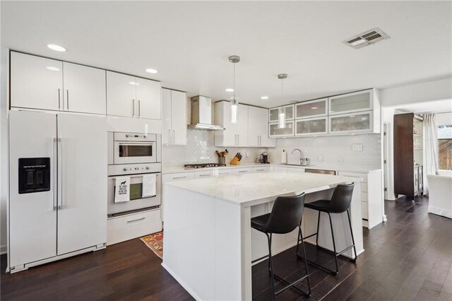 kitchen featuring white appliances, visible vents, wall chimney exhaust hood, glass insert cabinets, and dark wood-style flooring