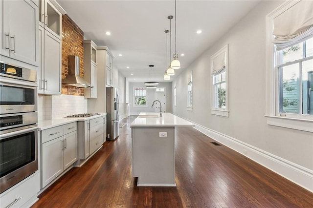 kitchen with gray cabinets, backsplash, appliances with stainless steel finishes, a sink, and wall chimney range hood