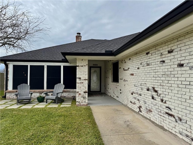 property entrance with brick siding, a lawn, a chimney, and roof with shingles