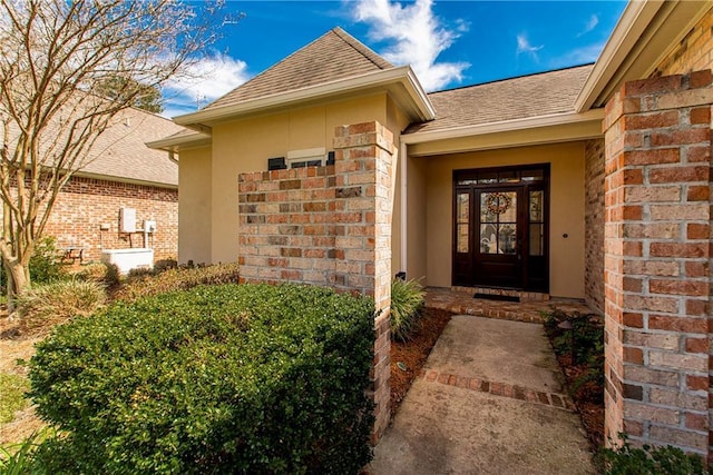 view of exterior entry with brick siding, a shingled roof, and stucco siding