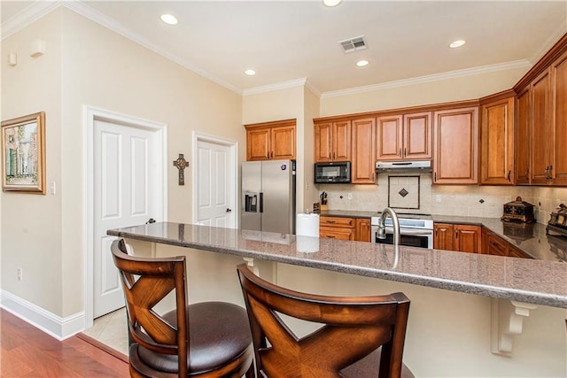 kitchen with a breakfast bar, brown cabinets, stainless steel appliances, visible vents, and under cabinet range hood