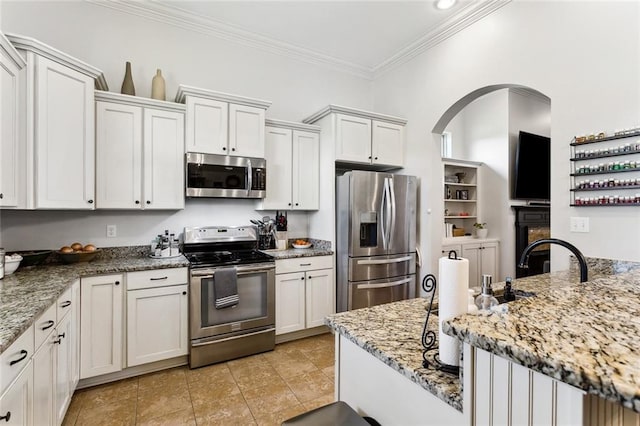 kitchen featuring light stone counters, arched walkways, white cabinets, appliances with stainless steel finishes, and crown molding