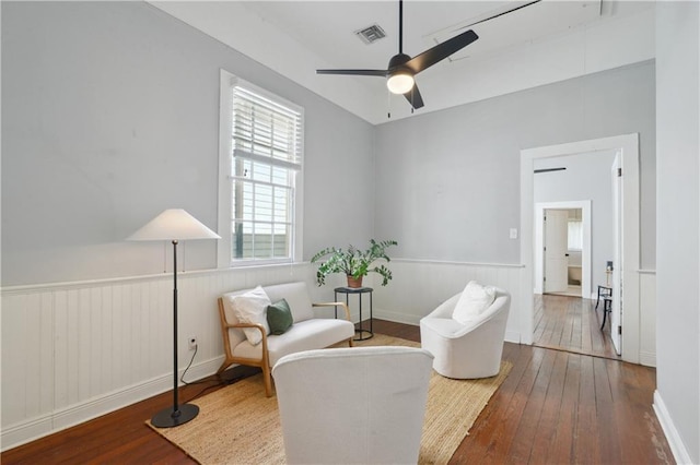 living area with a wainscoted wall, hardwood / wood-style flooring, visible vents, and ceiling fan