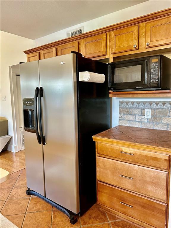 kitchen with visible vents, brown cabinets, stainless steel refrigerator with ice dispenser, tile countertops, and black microwave