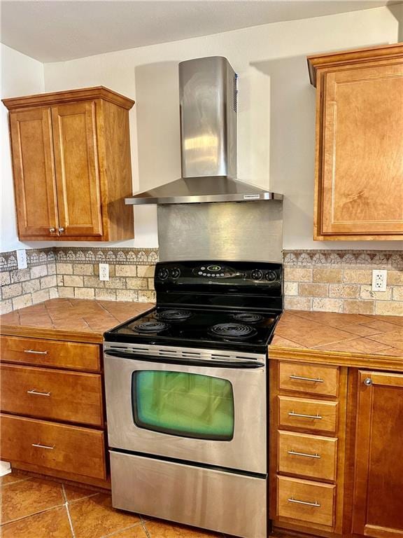 kitchen with wall chimney range hood, tile countertops, decorative backsplash, electric stove, and brown cabinetry