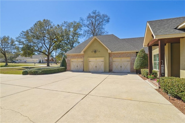 view of side of home featuring stucco siding, concrete driveway, an attached garage, a shingled roof, and brick siding