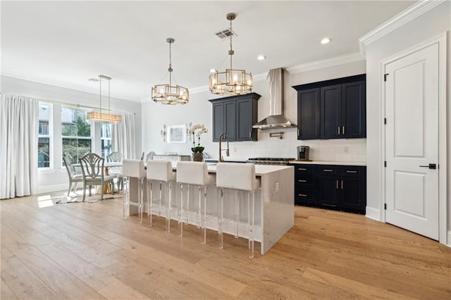 kitchen featuring tasteful backsplash, an island with sink, a sink, wall chimney exhaust hood, and dark cabinets