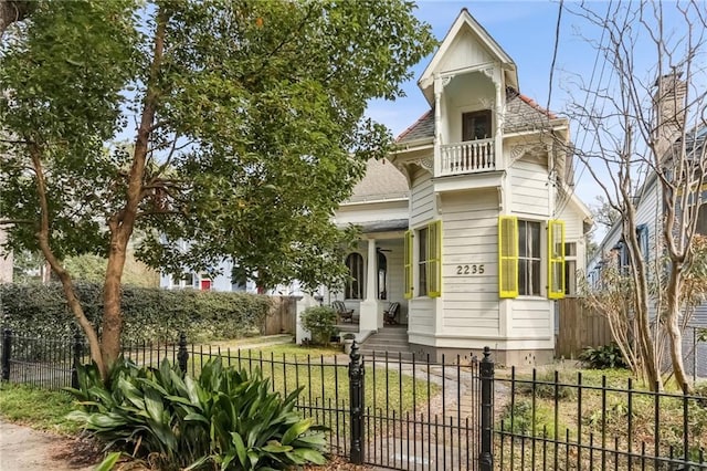 victorian house featuring a fenced front yard, a balcony, roof with shingles, and a gate