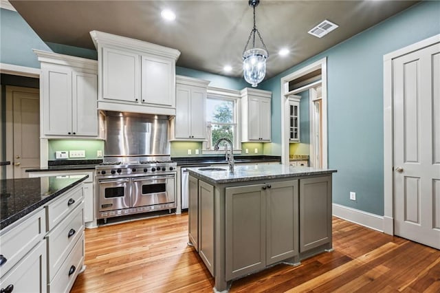 kitchen with visible vents, double oven range, dark stone counters, light wood-style flooring, and a kitchen island with sink